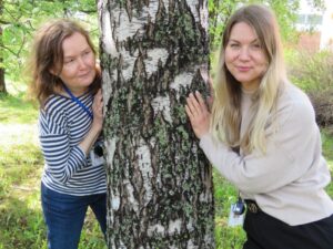 Osallisuuskoordinaattorit Heidi Snellman ja Sini Pulkkinen nojaamassa puunrunkoa vasten.