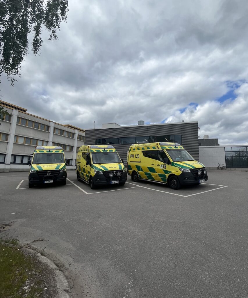 Kolme ambulanssia on pysäköitynä parkkiruutuihin. Taustalla on sairaala.
